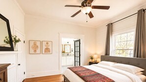 A bedroom with a bed, a ceiling fan, a window with curtains, a door leading to a bathroom, and a mirror with a vase of flowers. - Historic Craftsman, Walk to Duke (Durham)