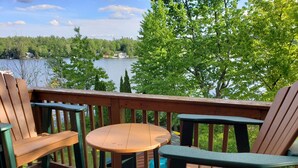 Eine Terrasse mit zwei Adirondack-Stühlen und einem kleinen runden Tisch mit Blick auf einen See, im Hintergrund Bäume und Häuser.