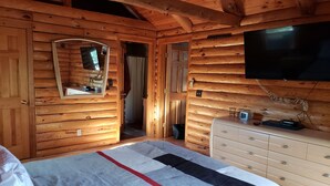 Ein Zimmer in einer Holzhütte mit einem Bett, einem Schminktisch, einem Fernseher und einem Spiegel.