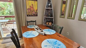 A wooden dining table with blue placemats, surrounded by chairs, in a room with a 'WELCOME TO OUR CABIN' sign, a wooden ladder shelf, and a view through a glass door.