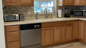 A kitchen with wooden cabinets, a dishwasher, a toaster, a microwave, and a window with a view of trees. - Venetian Vacation! Close To Beaches, Restaurants & Shopping! (Venice)