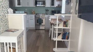 Cocina compacta con gabinetes blancos, horno empotrado y microondas. Hay un comedor con mesa y sillas blancas, y una estantería llena de libros.