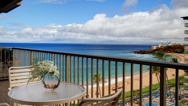 Een balkon met een tafel en een bloevas, met uitzicht op een strand met palmbomen en een helderblauwe lucht.