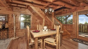 Interior de una cabaña de madera con una mesa de comedor, sillas, una escalera y vistas al exterior.