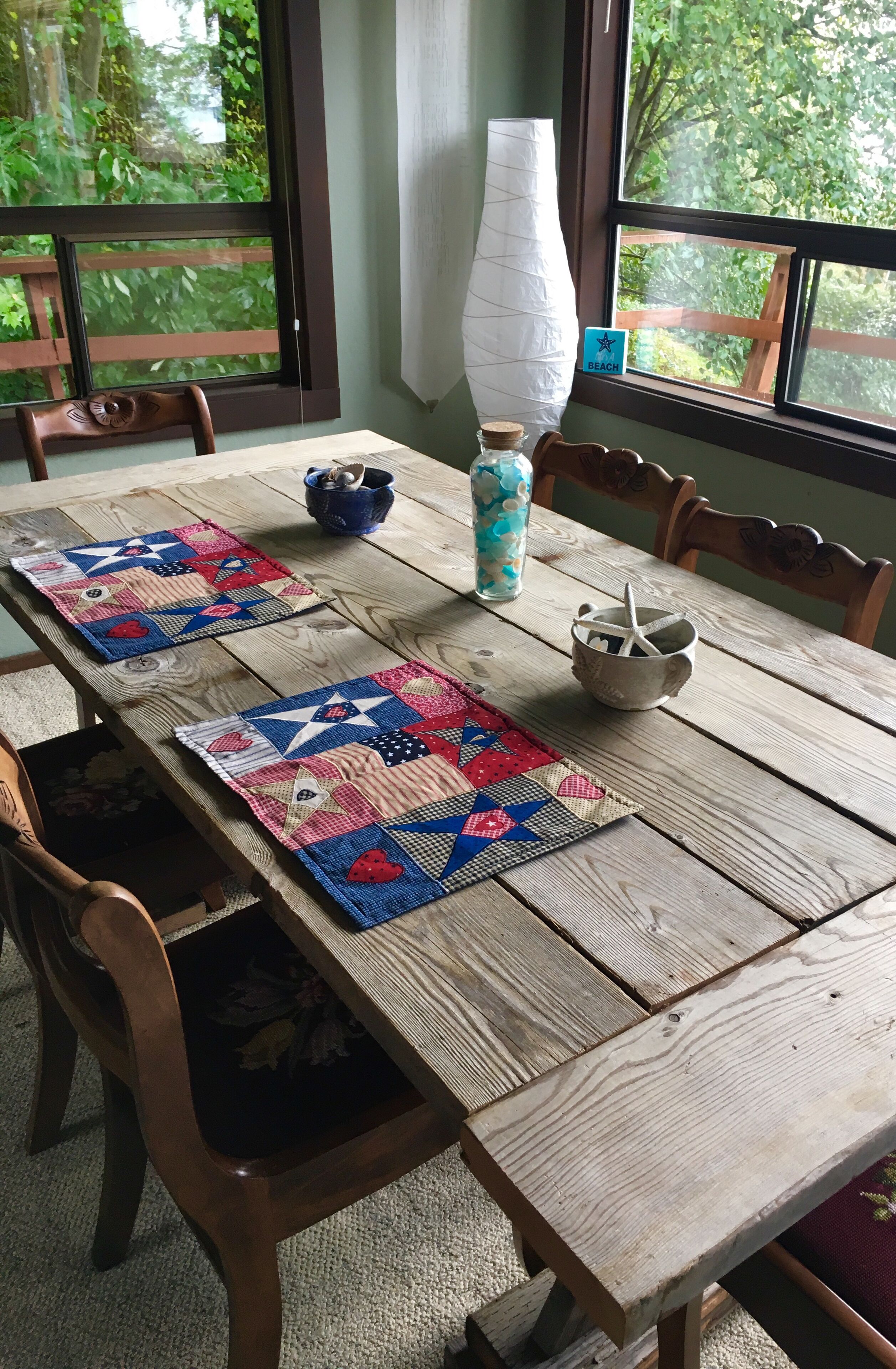 Une table à manger en bois, ornée de deux sets de table aux motifs d’étoiles et de cœurs, un grand vase blanc, une tasse bleue et un petit bol avec une cuillère.