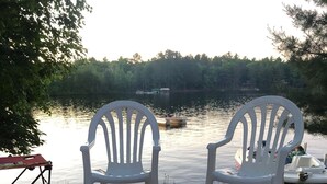 Un hogar con leña, dos sillas Adirondack blancas y un lago con barcos al fondo.