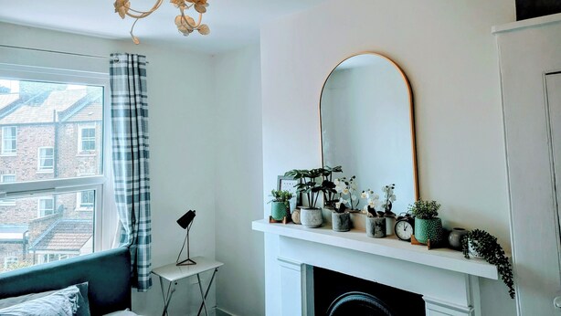 A bedroom with a bed, a window with curtains, a chandelier, a mirror, and a fireplace mantel with plants. - Luxury 4/5 bed 2 bath Victorian London home (London)