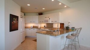 Eine moderne Küche mit weißen Schränken, einer Granit Arbeitsplatte und einer eingebauten Mikrowelle. Zwei Barhocker und eine kleine Kücheninsel mit Spüle sind ebenfalls vorhanden.
