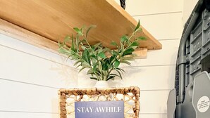A corner of a room with a wooden shelf holding a sign that reads "STAY AWHILE" and a potted plant.