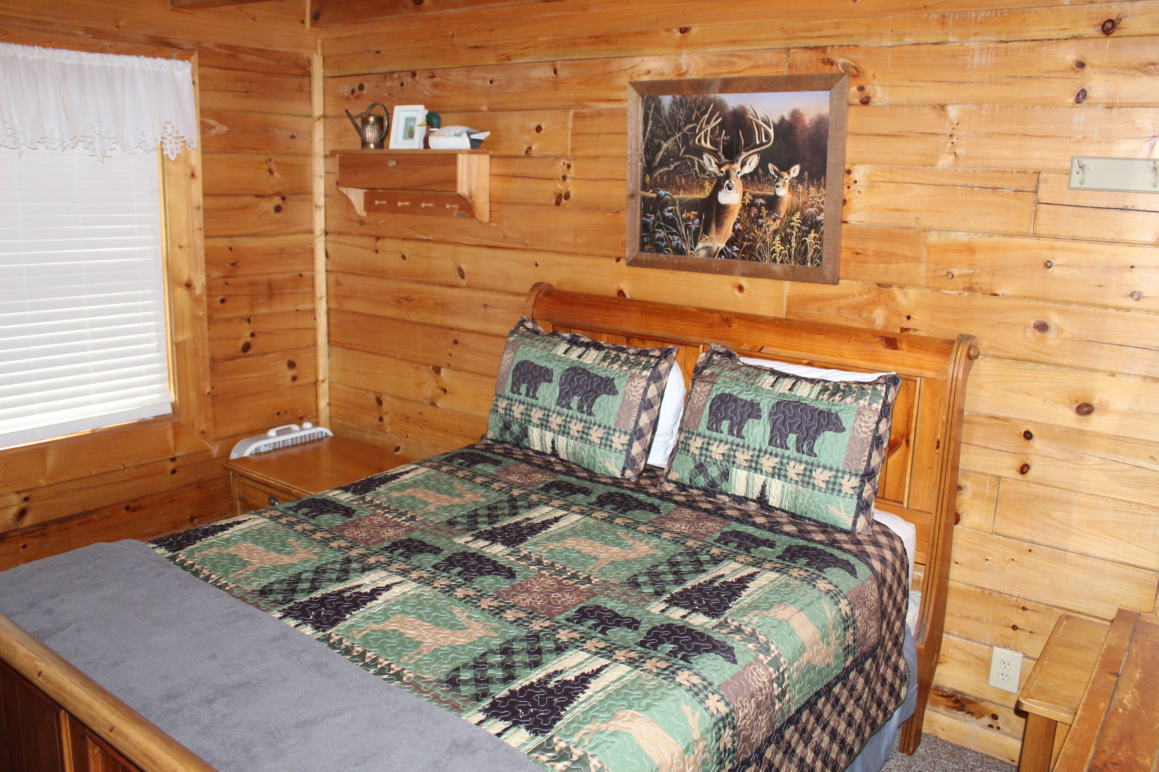 Ein Schlafzimmer einer Holzhütte mit einem Bett, einem Nachttisch, einer Ablage mit einem Bild und einem Fenster mit Jalousien.