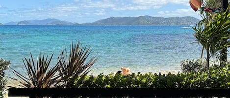 A view from a balcony overlooking a beach with clear blue water and distant mountains.