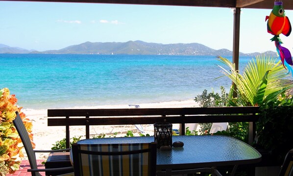 A beach view from a covered patio with a table and chairs, overlooking the sea and distant mountains.