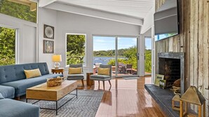 A spacious living room with a blue sofa, wooden coffee table, and a fireplace. Large windows offer a view of the outdoors. - Second Wind :039-O  An Orleans Estate with large heated pool, spa, on Town Cove; sleeps 24 (Orleans)
