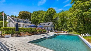 A swimming pool with lounge chairs and umbrellas, surrounded by a landscaped garden and a large building in the background. - Second Wind :039-O  An Orleans Estate with large heated pool, spa, on Town Cove; sleeps 24 (Orleans)