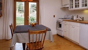 Zona pranzo con tavolo apparecchiato per due, una sedia di legno, una ciotola di frutta e vista su un giardino attraverso una porta in vetro.