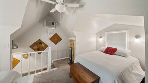 A bedroom with a sloped ceiling, a ceiling fan, a bed with white bedding, a wooden bench, a desk with a lamp, and a wall-mounted heater. - Pool Side Guest House On Lake Hudson Near Pryor (Adair)