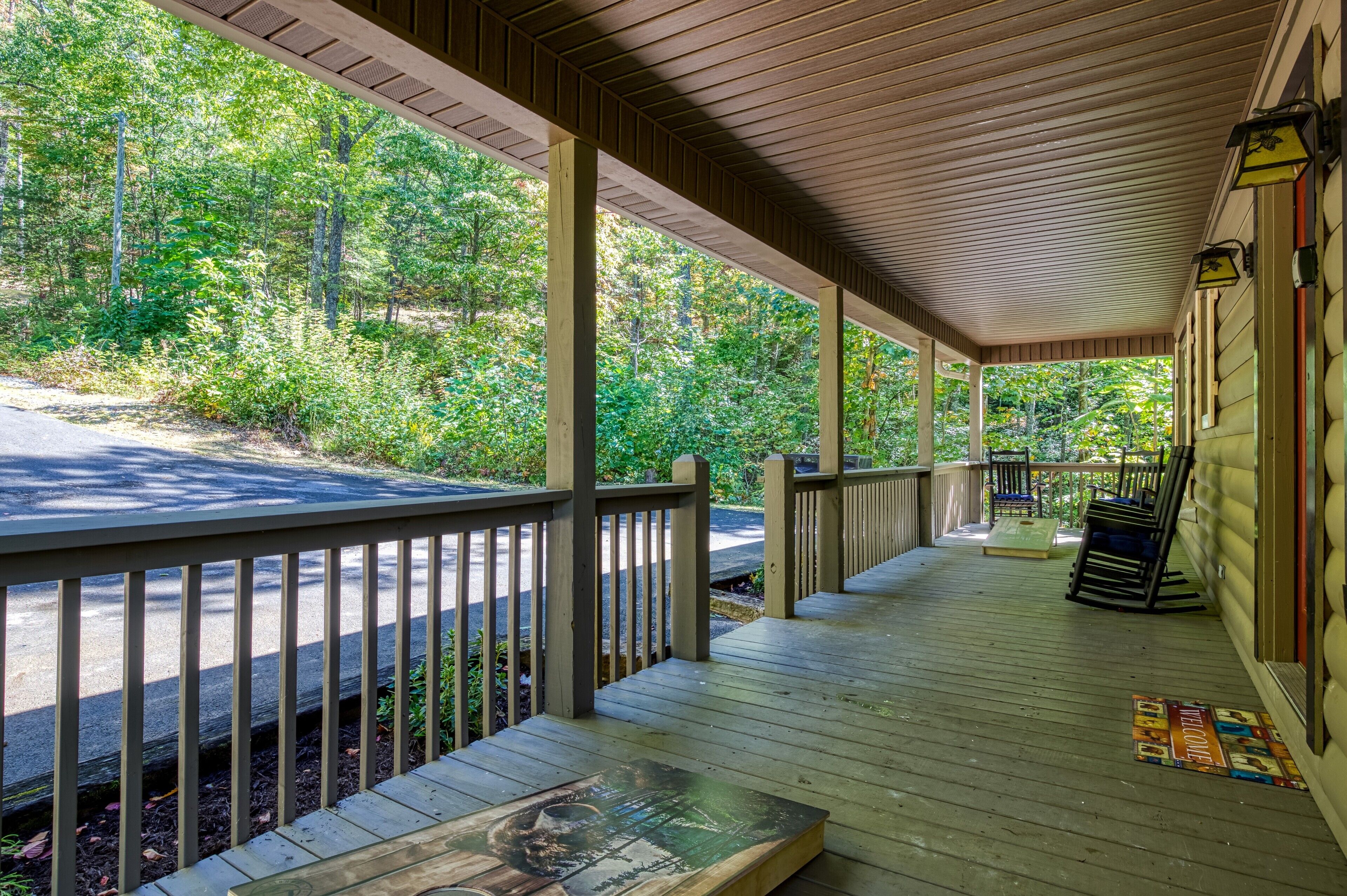 A covered porch with a wooden deck, railings, and a view of a forested area.