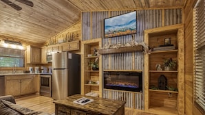 Un intérieur rustique en bois, avec une cuisine, une cheminée et un téléviseur fixé au mur.