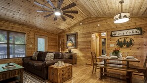 Interior acogedor de una cabaña con techo de madera, ventilador de techo, mesa de comedor con platos y vasos, sofá de cuero marrón y cómoda de madera.