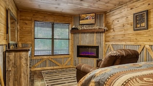 Una habitación rústica de madera con un ventanal grande, una chimenea, un sillón de cuero y una cama con una colcha estampada.