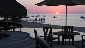 A beachside seating area with a table and chairs, a thatched umbrella, and a view of the sunset over the sea with boats. - VILLAS DE CORTEZ, Beachfront, 24 PICKLEBBALL CTS, Poolside, ,  1 BR Condo,  (Los Barriles)