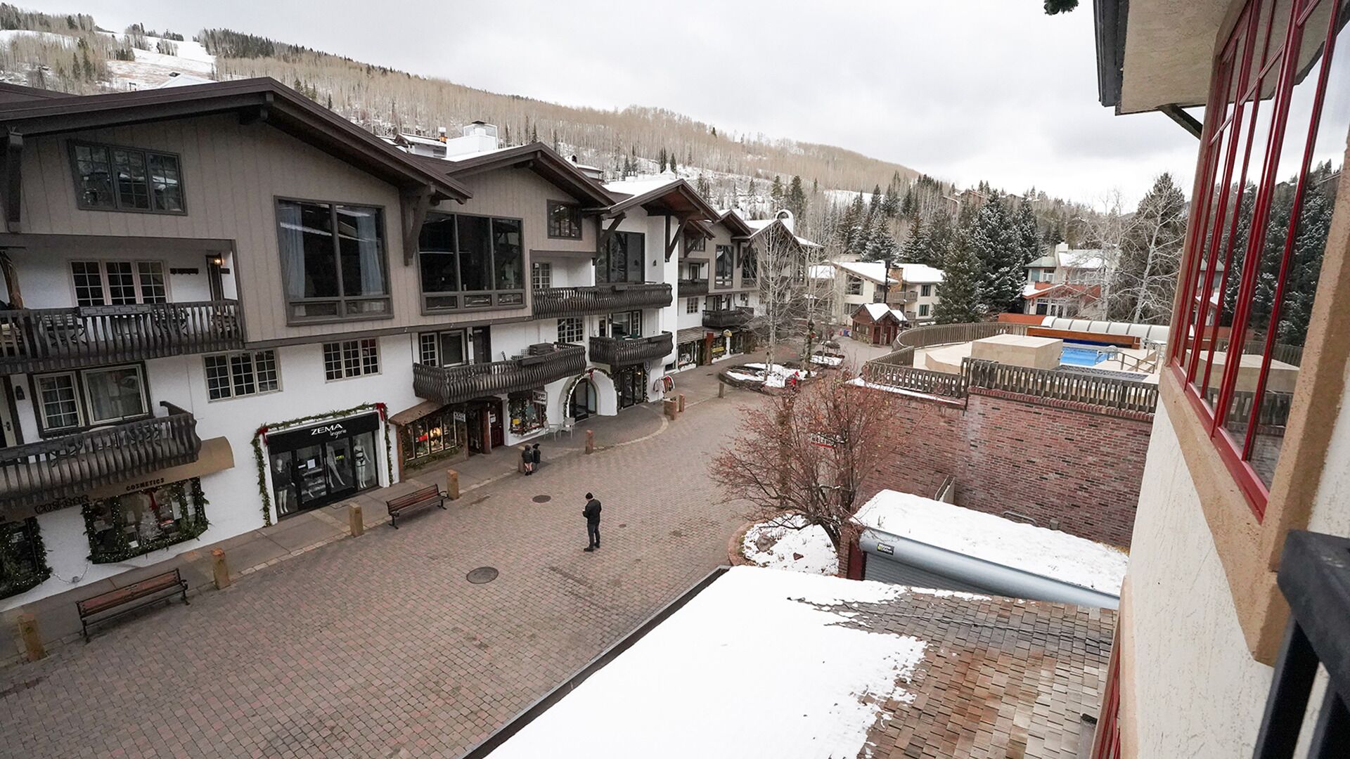 Une cour d’hôtel avec un chemin pavé, des bâtiments à plusieurs étages dotés de balcons, et une chaîne de montagnes enneigées en arrière-plan.