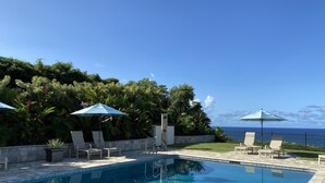 Una piscina con sillas de descanso y sombrillas, rodeada de exuberante vegetación y vistas al océano.