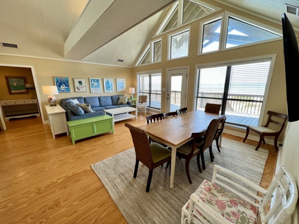 A spacious living area with a wooden dining table, comfortable seating, a green sofa, and large windows providing natural light.