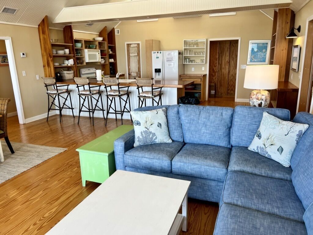 A living room with a blue sofa, a green bench, and a white table. There is a kitchen area in the background with a refrigerator, microwave, and cabinets.