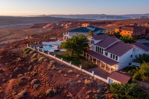 Ein großes, modernes Haus mit Swimmingpool, umgeben von einem weißen Zaun, in einer Wüstenlandschaft mit Bergen im Hintergrund.