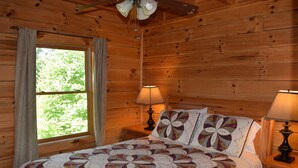 Un dormitorio de cabaña de madera con una cama, dos lámparas de noche, una ventana con cortinas y un ventilador de techo.