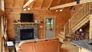 Interior de una cabaña de troncos con chimenea de piedra, un televisor montado encima, un sofá y una escalera que lleva a un nivel superior.