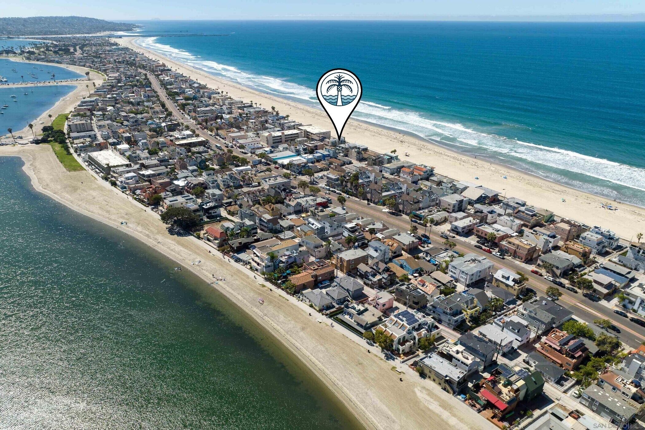 An aerial view of a coastal residential area with houses along the beachfront, a clear view of the ocean, and a beach with people and boats.