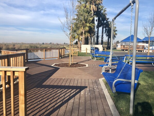 Una terraza de madera con bancos y un columpio, rodeada de palmeras y una valla.