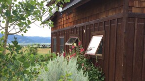 A rustic wooden building with a sloped roof, surrounded by lush greenery and flowering plants. - Sweet and charming Victor home on three acres with Teton views! (Victor)