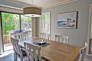 Een eetruimte met een houten tafel, witte stoelen, een geweven hanglamp en een schilderij van een kustlandschap in een lijst.