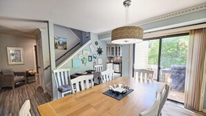 Un coin repas avec une table en bois, des chaises blanches, un luminaire suspendu et une vue sur l’extérieur grâce à une porte-fenêtre coulissante.