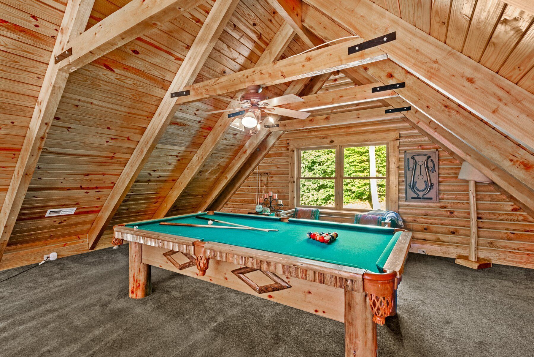 Une pièce mansardée en bois, équipée d’une table de billard, d’un ventilateur de plafond et d’une fenêtre donnant sur un paysage verdoyant.