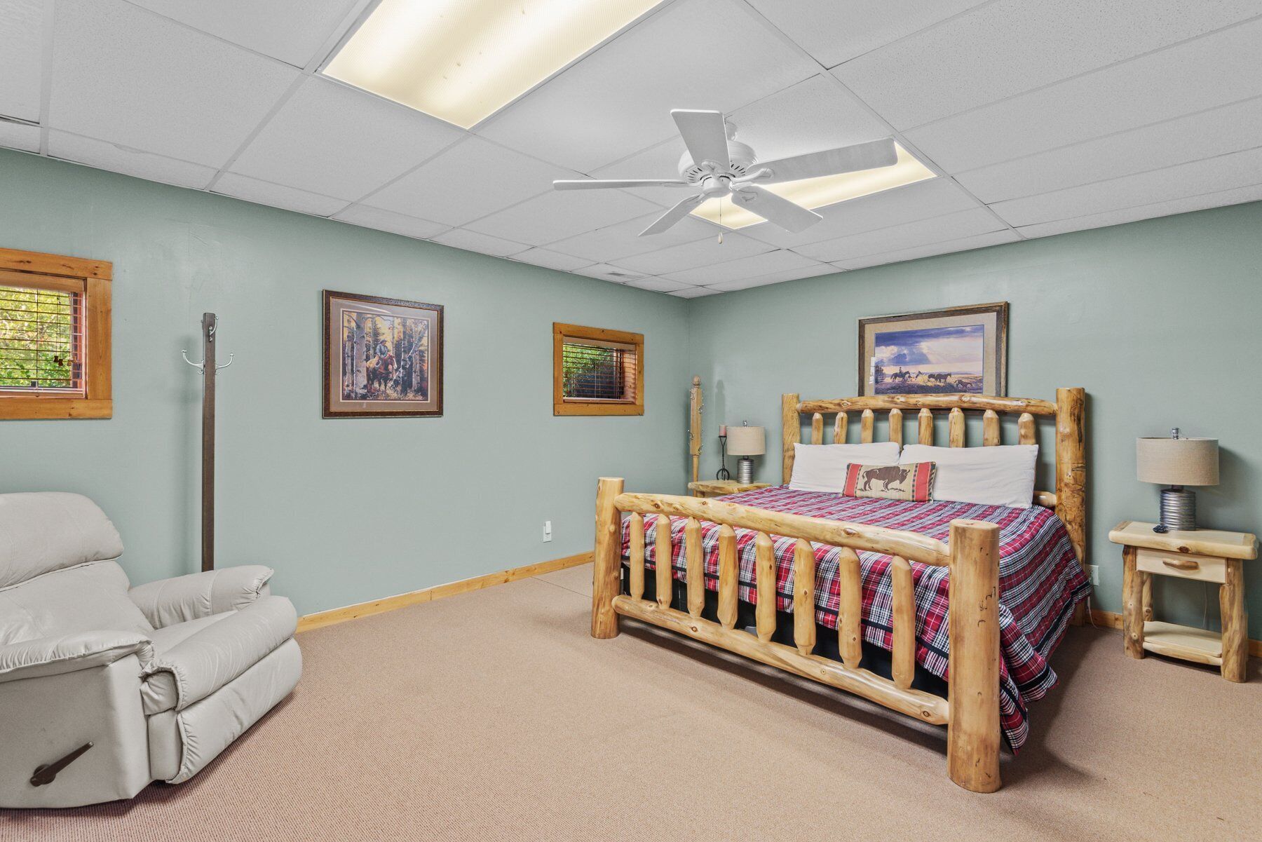 A bedroom with a wooden bed, a reclining chair, a ceiling fan, and framed pictures on the walls.