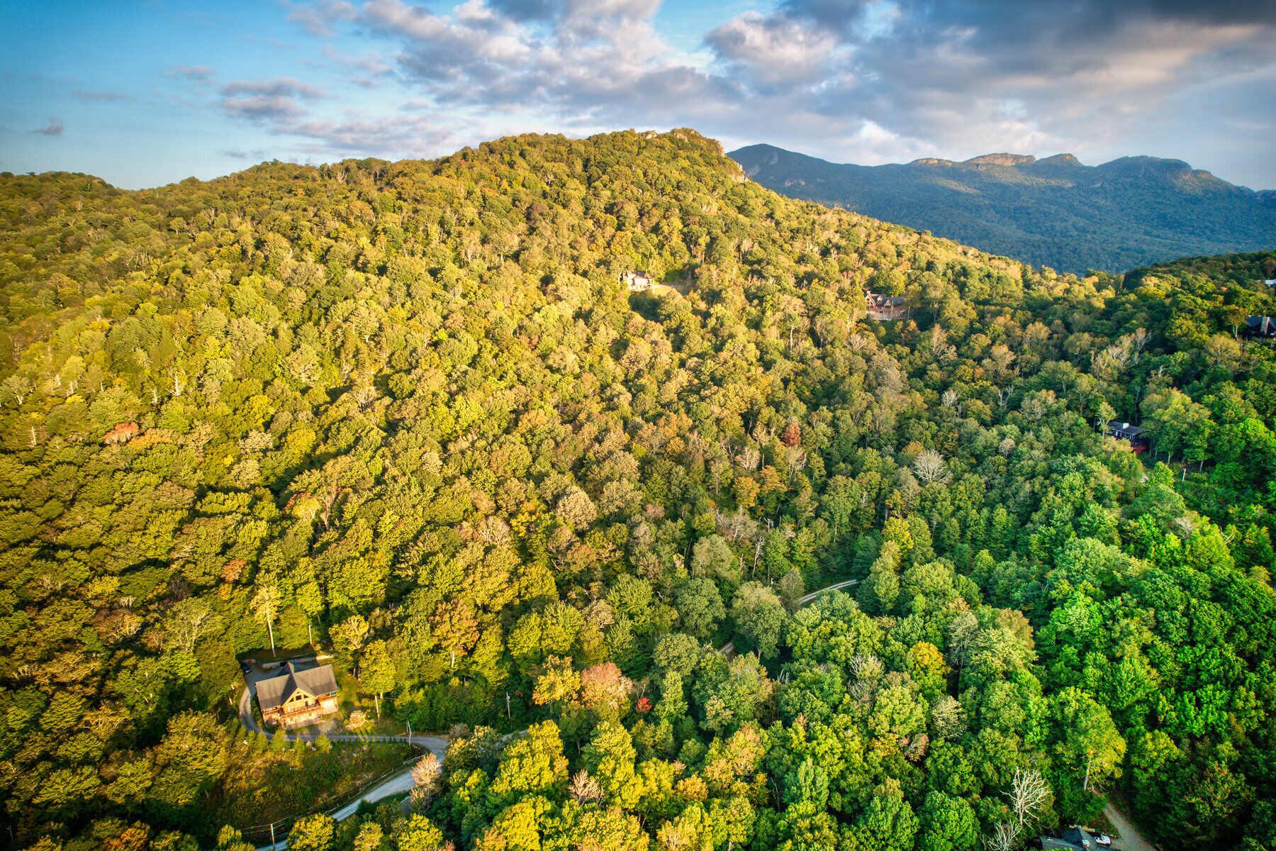 Vista aérea de una zona montañosa cubierta de bosques, con varias construcciones, incluyendo una casa con un camino de entrada visible y una pequeña edificación cerca de una carretera.