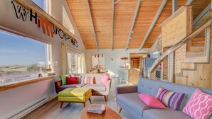 A cozy interior with a wooden ceiling, a large window offering a view, and a colorful sofa with cushions. - Whimsical Ocean Front Home! Hot tub & Dog Friendly! (Waldport)