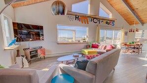 A spacious living room with a high ceiling, a large window, and a view of the outdoors. There is a sofa, a coffee table, and a dining area with chairs. - Whimsical Ocean Front Home! Hot tub & Dog Friendly! (Waldport)