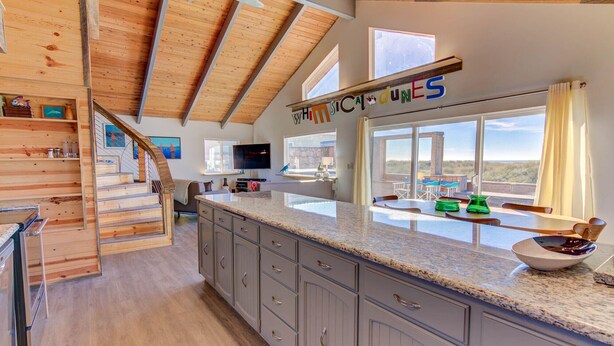 A spacious kitchen with a large island, wooden beams on the ceiling, and a view of the outdoors through large windows. - Whimsical Ocean Front Home! Hot tub & Dog Friendly! (Waldport)