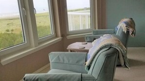 Een kamer met uitzicht op het strand door grote ramen, een blauwe fauteuil en een klein tafeltje met een plaid.