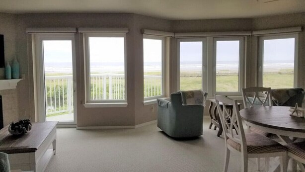 Ruime kamer met grote ramen en uitzicht op het strand. Een eettafel met stoelen en een comfortabele zithoek.