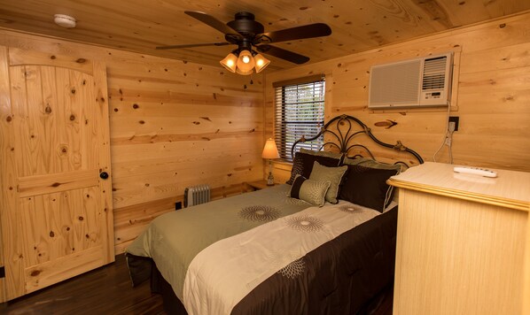 A wooden cabin bedroom with a bed, a ceiling fan, a window with blinds, and a wall-mounted air conditioner. - WAE Outdoors Cabin # 4 Log Cabin (Cliffton)