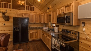 A wooden cabin interior with a black refrigerator, stainless steel microwave, oven, and a wooden deer head mounted on the wall.
