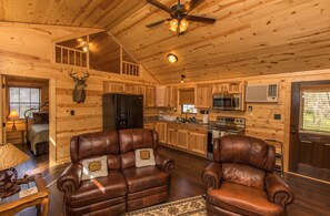 A cabin interior with a wooden interior, a ceiling fan, a kitchen area, and leather furniture. - WAE Outdoors Cabin # 4 Log Cabin (Cliffton)