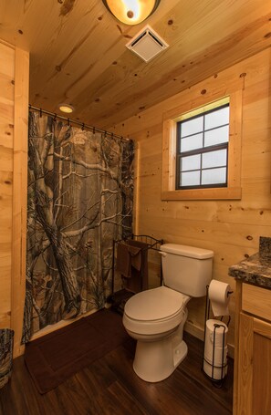 A wooden cabin bathroom with a camouflage shower curtain, a toilet, a window, and a marble countertop. - WAE Outdoors Cabin # 4 Log Cabin (Cliffton)