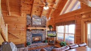 Un acogedor interior de cabaña con chimenea de piedra, sofá de cuero y televisor sobre un soporte de madera.
